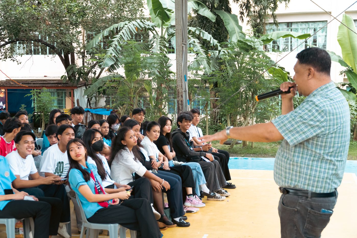 A speaker holding a microphone gestures toward rows of seated students, several of whom are smiling.