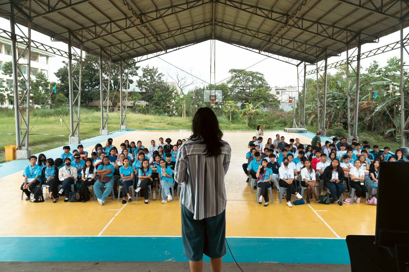 Seen from behind a speaker on a covered outdoor court, rows of students in blue uniforms sit facing the stage.