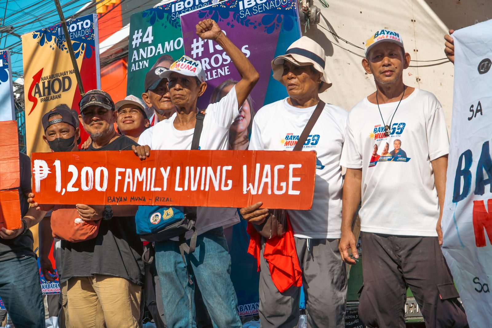 Workers stand behind a hand-painted orange placard reading P1,200 family living wage. One raises a clenched fist. Senatorial campaign banners hang behind them in the sun.
