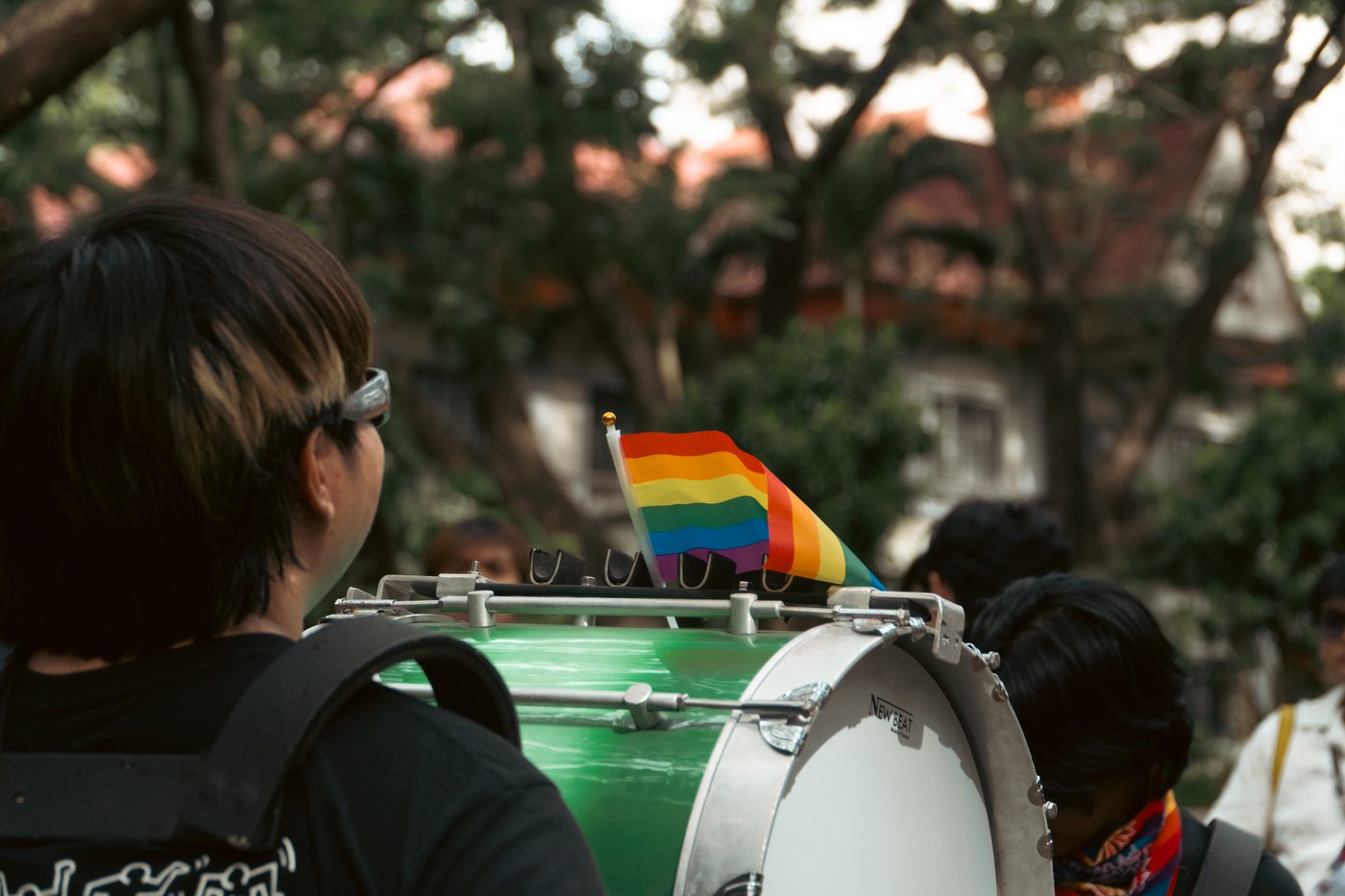 A drummer carries a green bass drum with a small rainbow flag fixed to the rim, seen from behind in warm afternoon light.