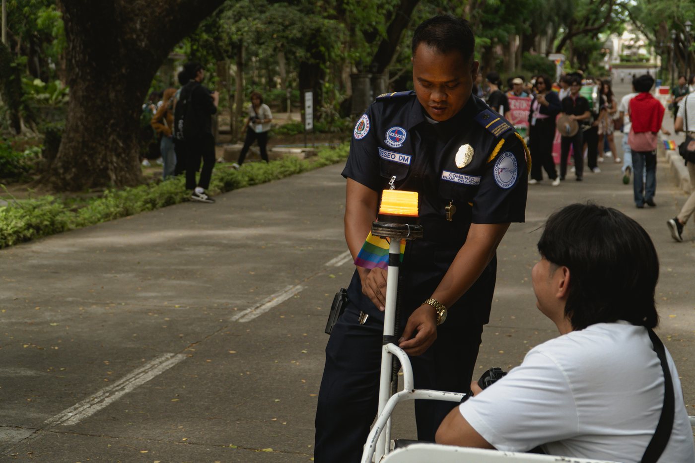 A campus security guard leans on a wheeled barrier post wrapped in a rainbow flag, talking with someone seated at the roadside. Marchers move along the tree-lined road behind.