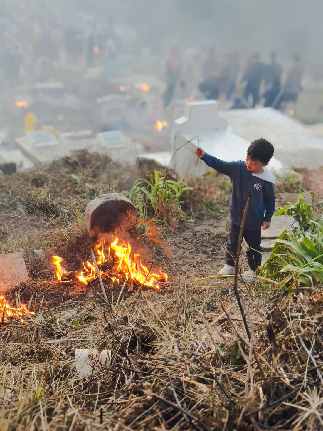 A small boy in a navy jacket holds a length of stick out towards a low fire burning on the ground beside a grave.