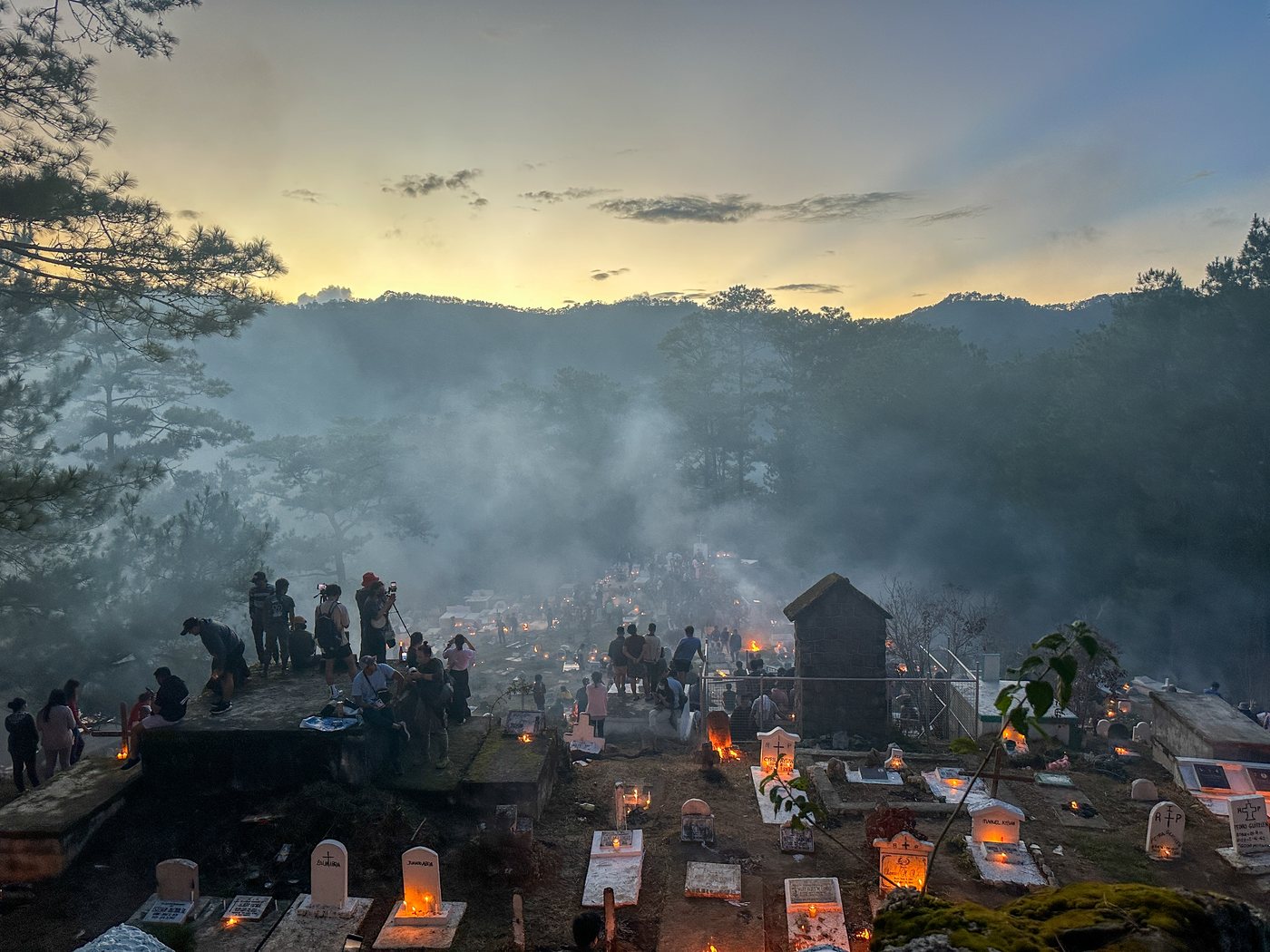 Blue hour over a hillside cemetery. Small fires and candles burn on the graves, smoke drifts through pine trees, and a group of photographers stands on a tomb at the left edge.