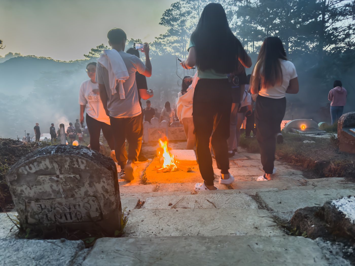 People walk single file along a stone path between graves, passing a fire that lights the ground orange. Smoke fills the valley behind them.