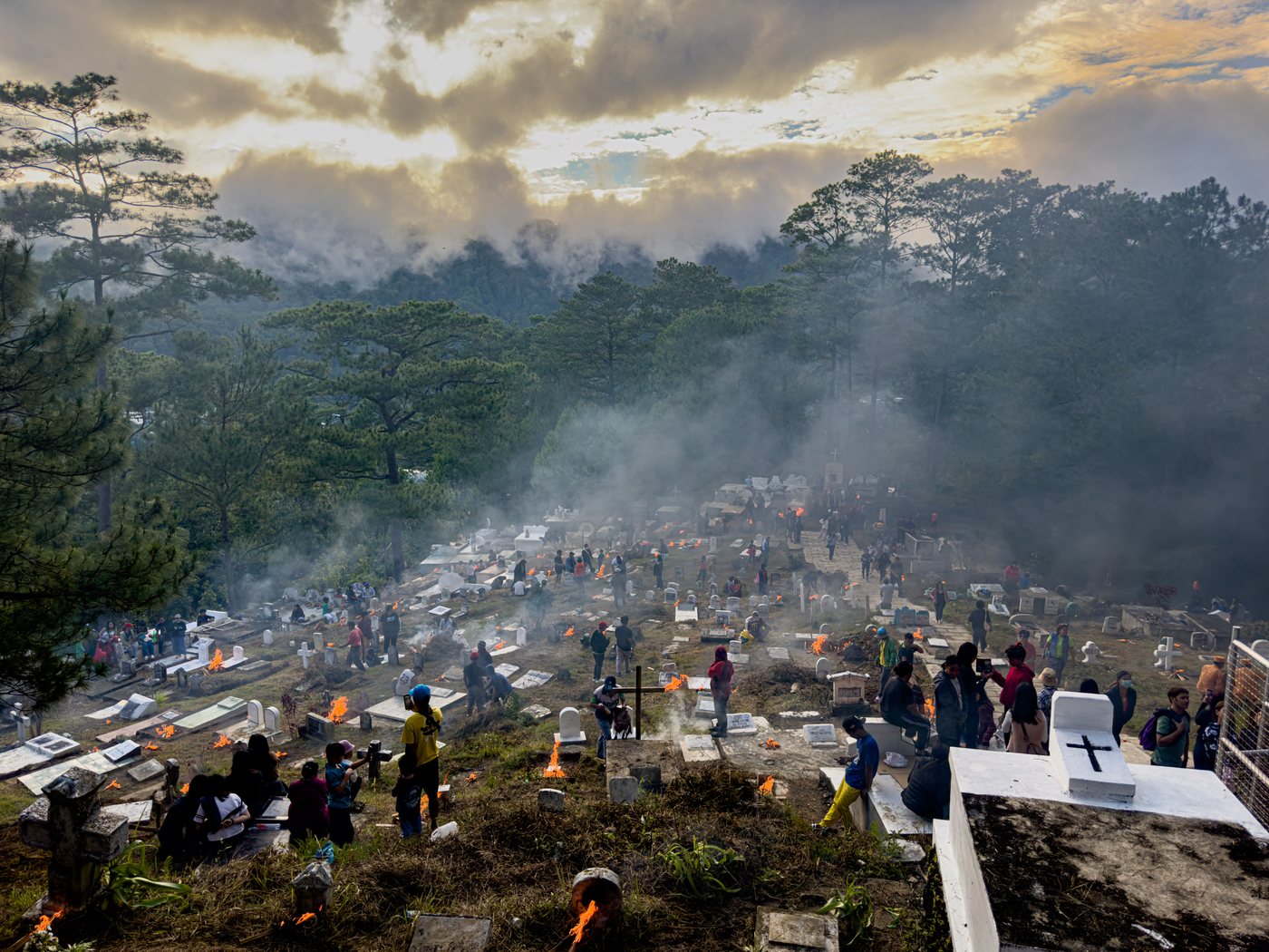 Elevated view across the cemetery. Dozens of small fires burn among the headstones and smoke layers across the pine forest under a bright evening sky.
